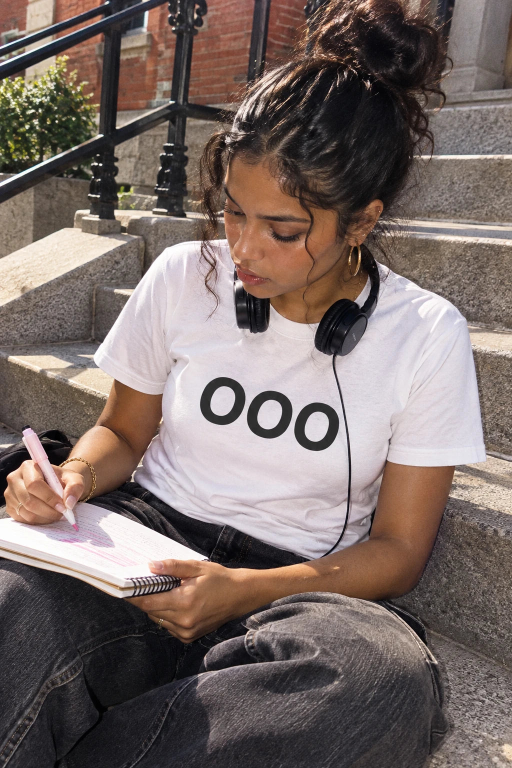 Woman wearing a white OOO T-shirt and headphones while writing in a notebook on outdoor steps.