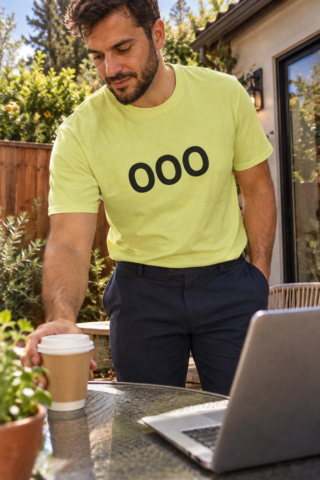Man wearing a pale yellow-green OOO T-shirt while setting coffee beside a laptop on a garden patio.