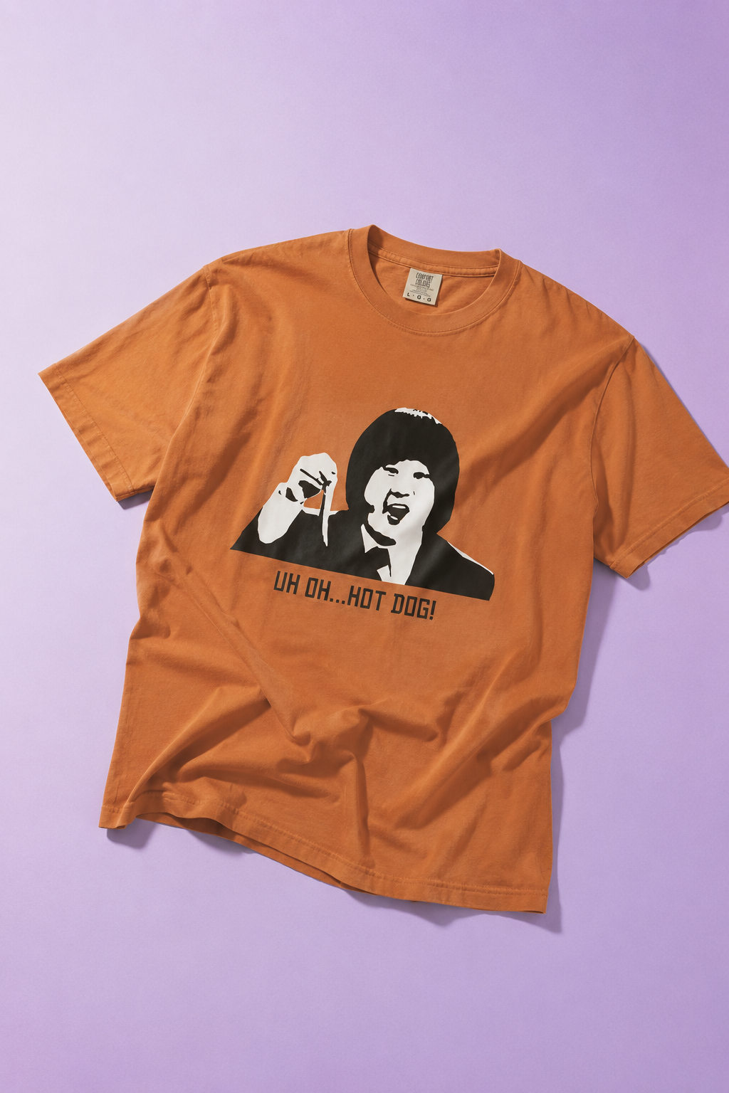 Orange graphic T-shirt laid flat on a light purple background featuring a black-and-white portrait print with the text "UH OH... HOT DOG!"
