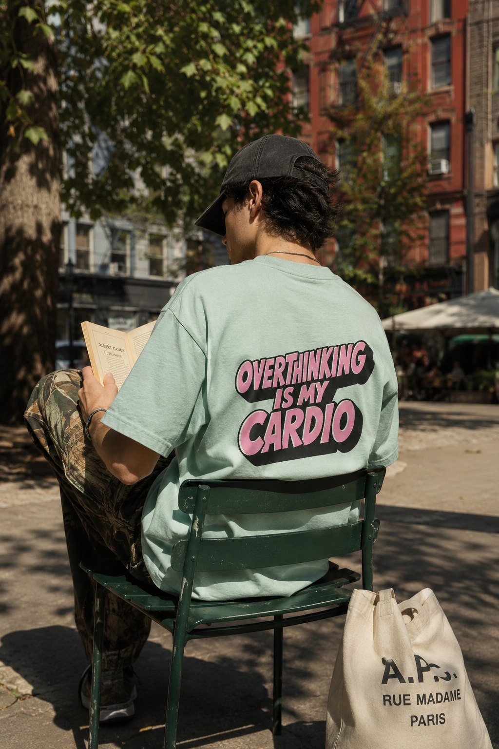 Back view of model wearing sage green Overthinking Is My Cardio graphic T-shirt outdoors