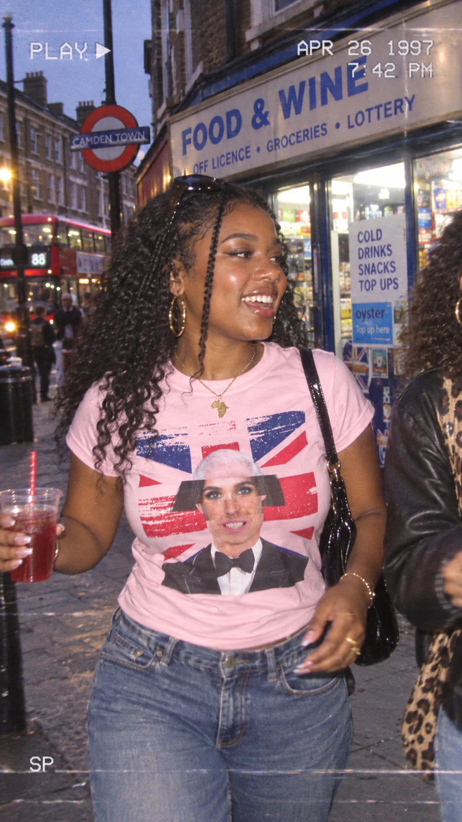 Woman wearing the light pink Tantrum Media Royal Frankenstein T-Shirt outside a food and wine shop at night.