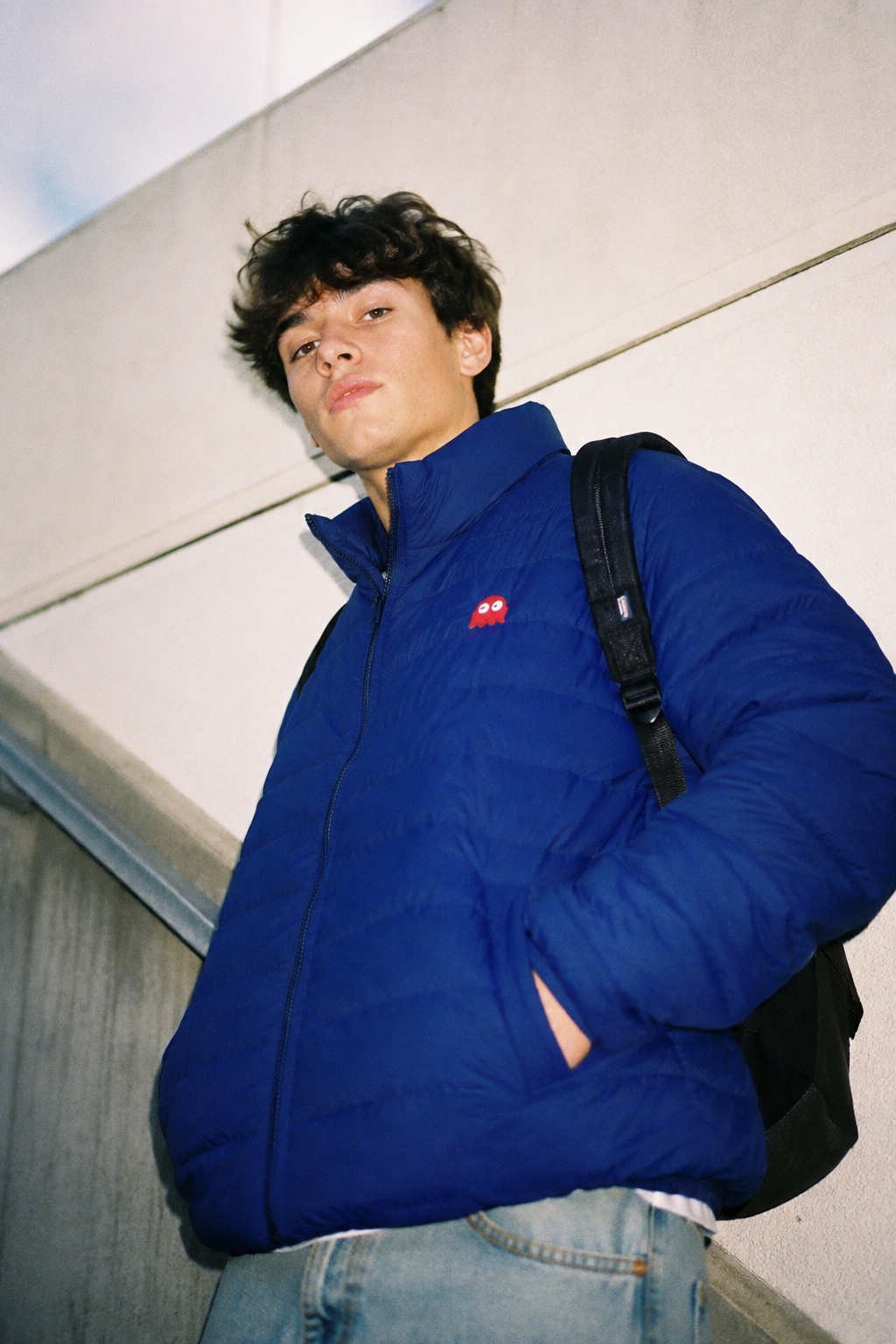Gen Z student wearing the Pac-Man Blinky Ghost men’s blue puffer jacket against a light college backdrop in a dramatic low-angle lifestyle shot.