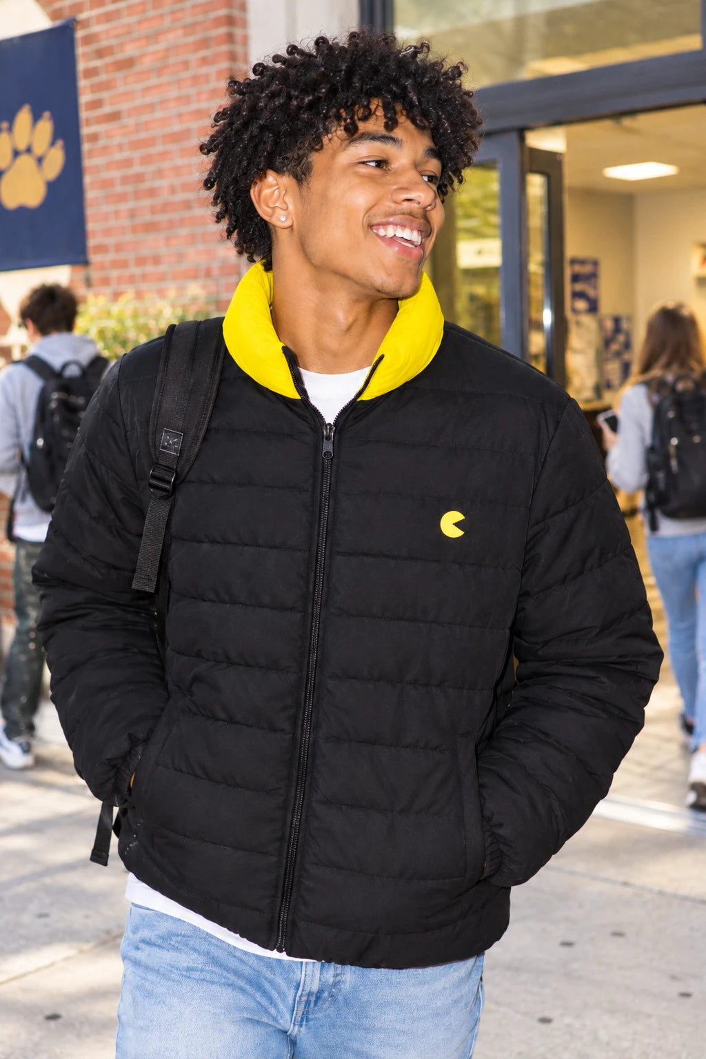Gen Z man wearing Pac-Man men’s black and yellow puffer jacket with yellow collar and chest logo on a college campus