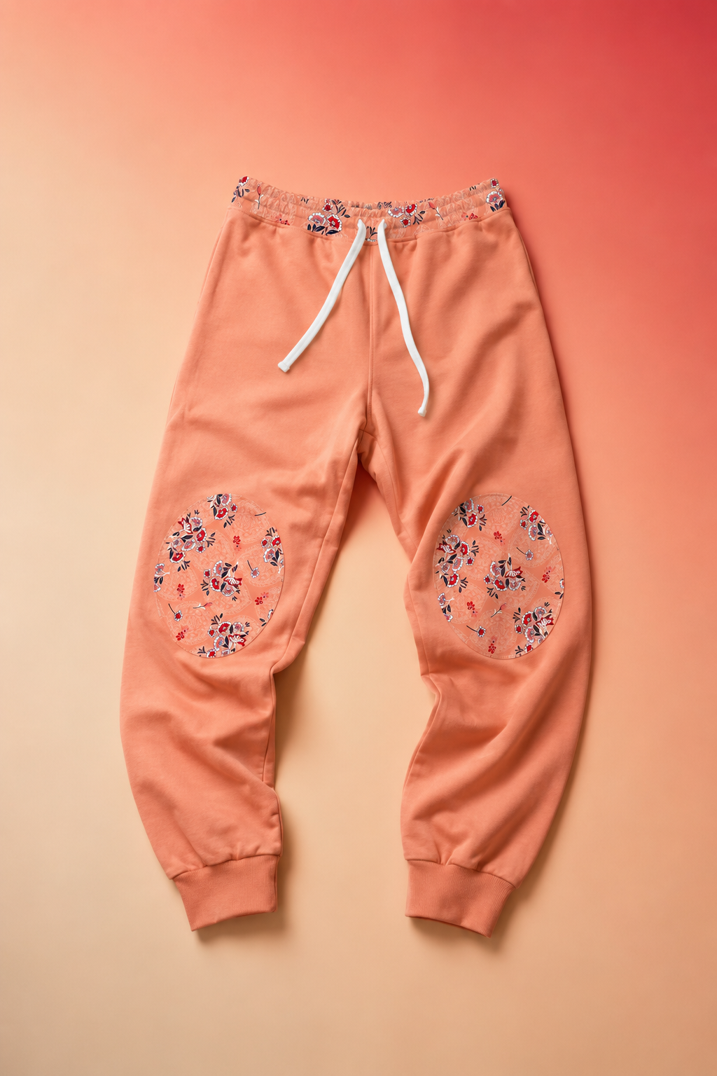 Peach floral joggers displayed on a soft coral gradient background in an editorial flat lay