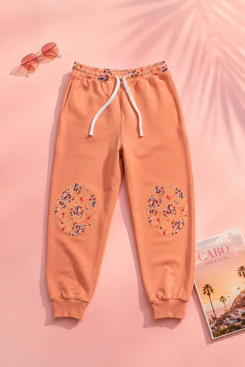 Peach floral joggers with floral knee patch details styled on a soft pink editorial flat lay background