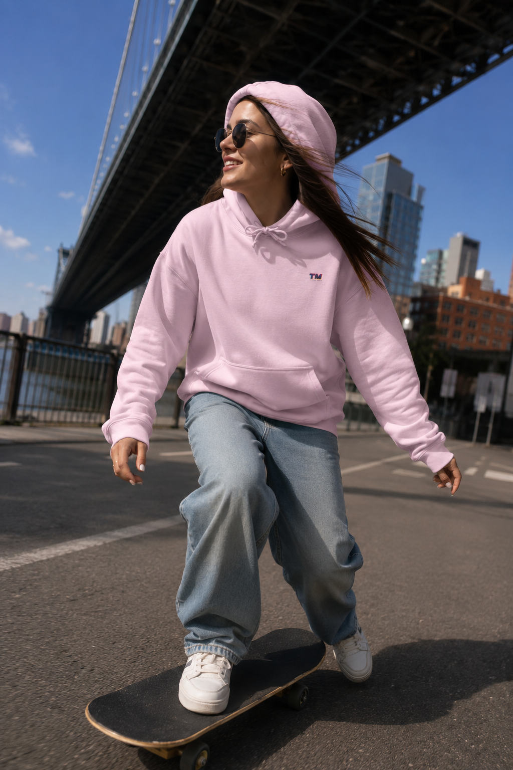 Female Gen Z skateboarding beneath the Brooklyn Bridge wearing the Tantrum Media EcoSmart pink hoodie.