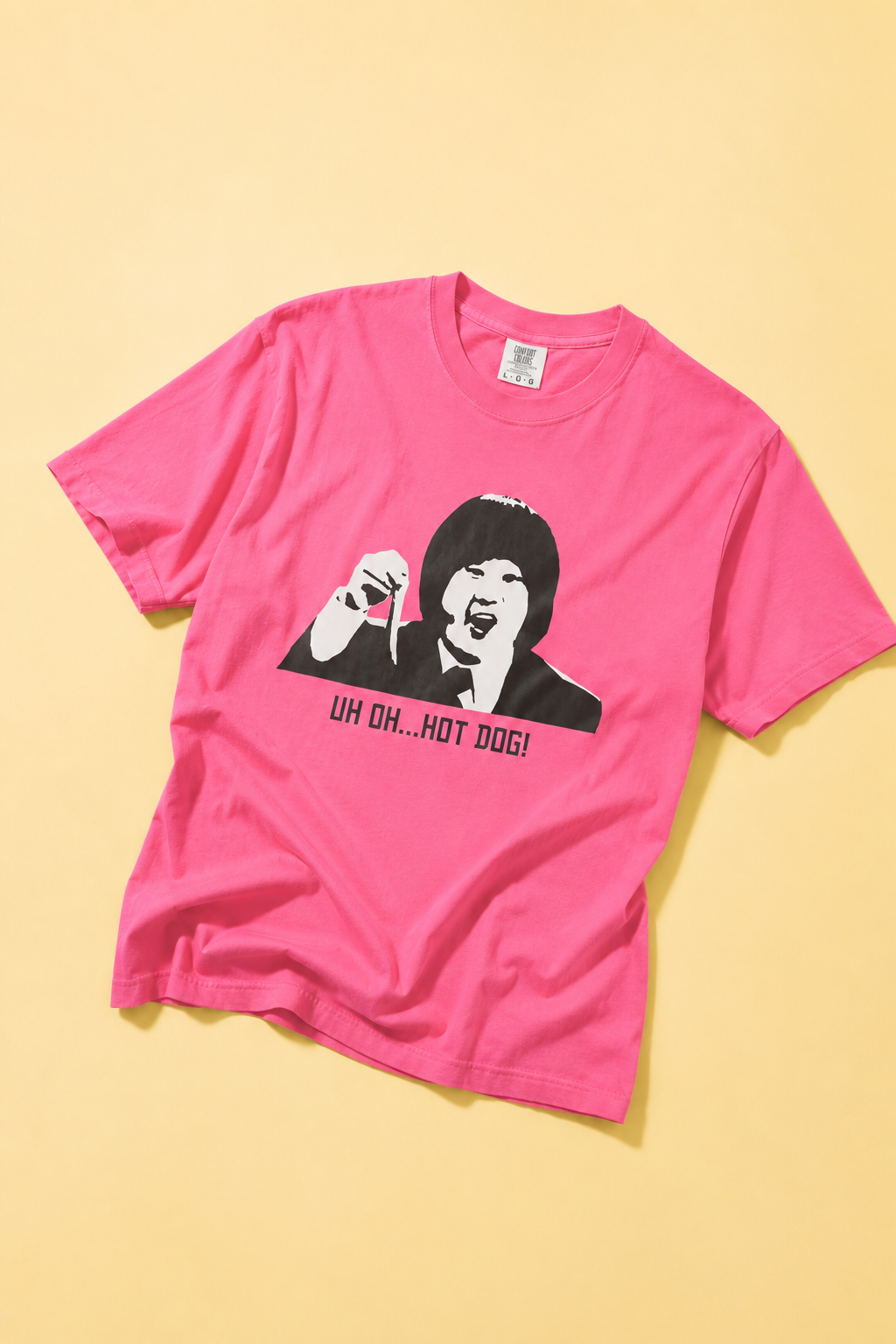 Bright pink graphic T-shirt laid flat on a yellow background featuring a black-and-white portrait print with the text "UH OH... HOT DOG!"
