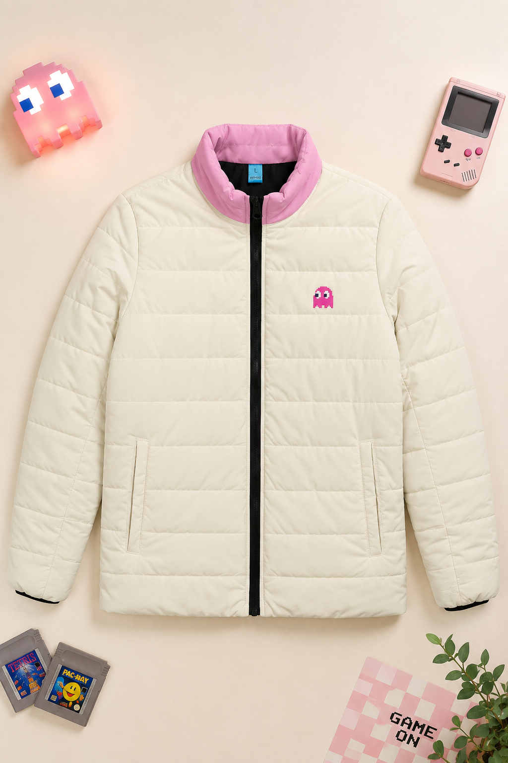 A cream-colored puffer jacket styled in a flat-lay with retro gaming props, featuring a pink collar, black zipper, and a pink Pac-Man logo on the chest.