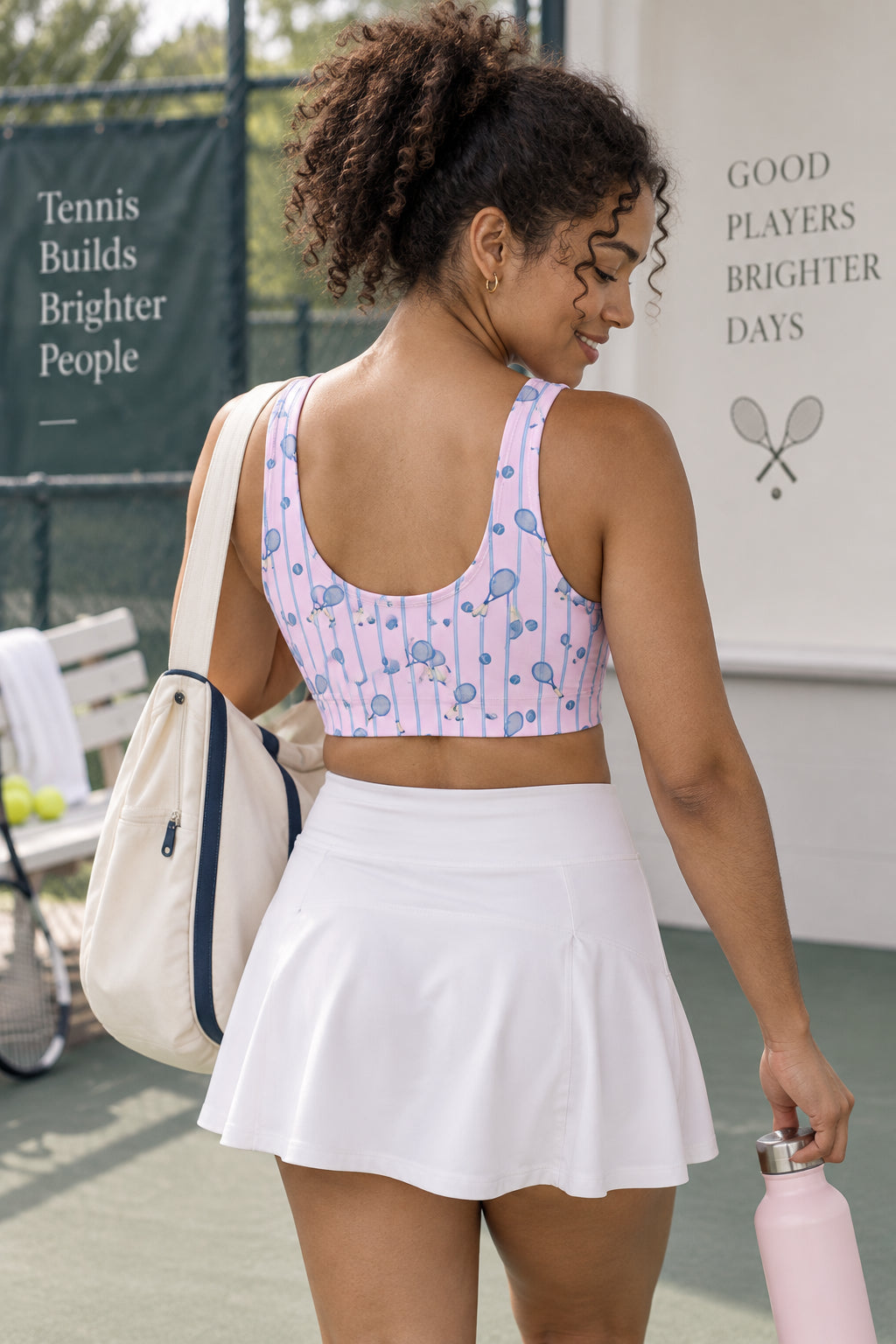 Back view of woman wearing a pink tennis print sports bra with a U-shaped back and white tennis skirt while carrying a tennis bag and water bottle.