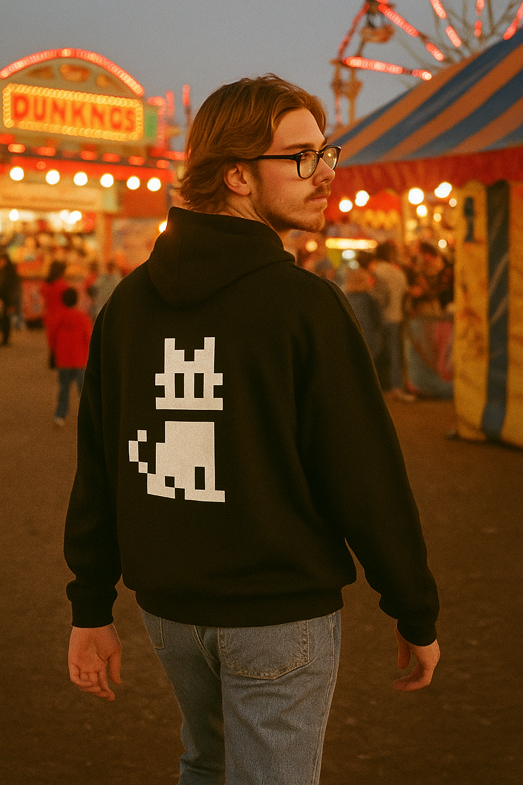 Person standing at an outdoor fair in the evening wearing a black hoodie with a large white pixel cat graphic on the back, colorful carnival lights and booths blurred in the background.