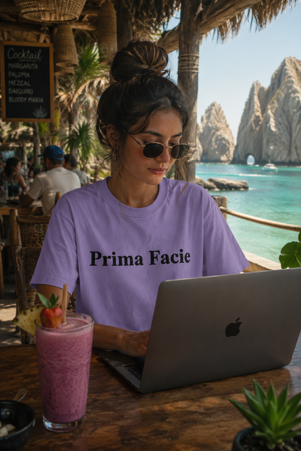 Female Gen Z wearing a lavender Prima Facie graphic T-shirt while working on a laptop and drinking a smoothie at a beach bar in Cabo San Lucas with cinematic tropical lighting and ocean cliffs in the background.