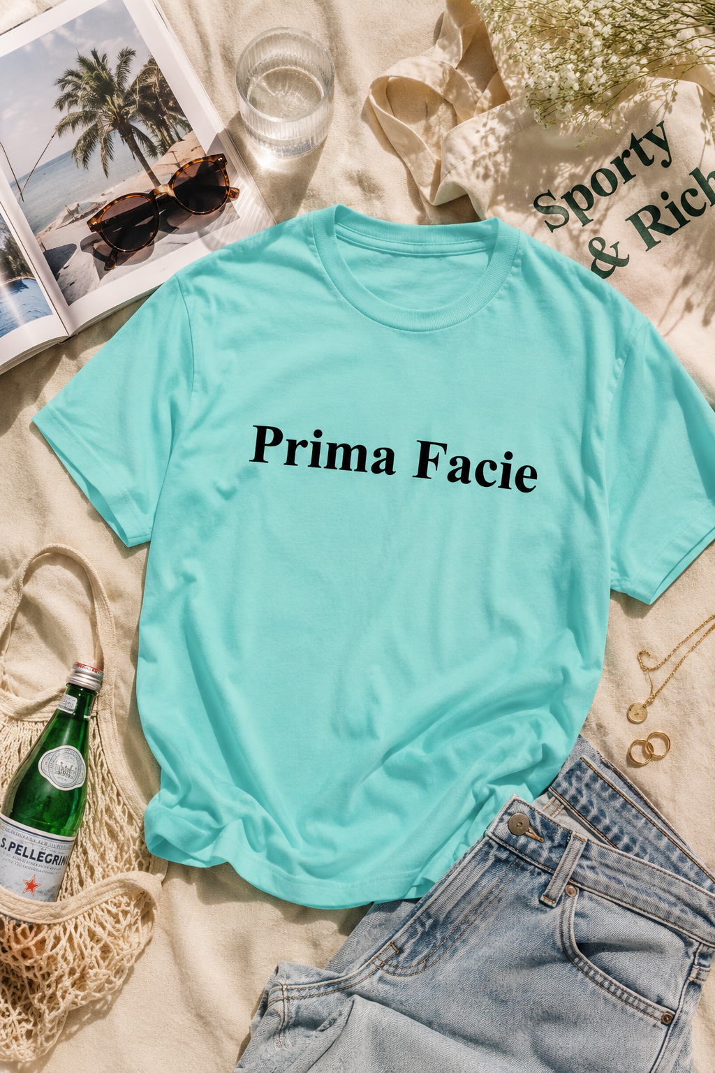Editorial flat lay of a mint Prima Facie graphic T-shirt styled with denim jeans, sunglasses, jewelry, tote bag, sparkling water bottle, tropical magazine, and beach-inspired lighting.