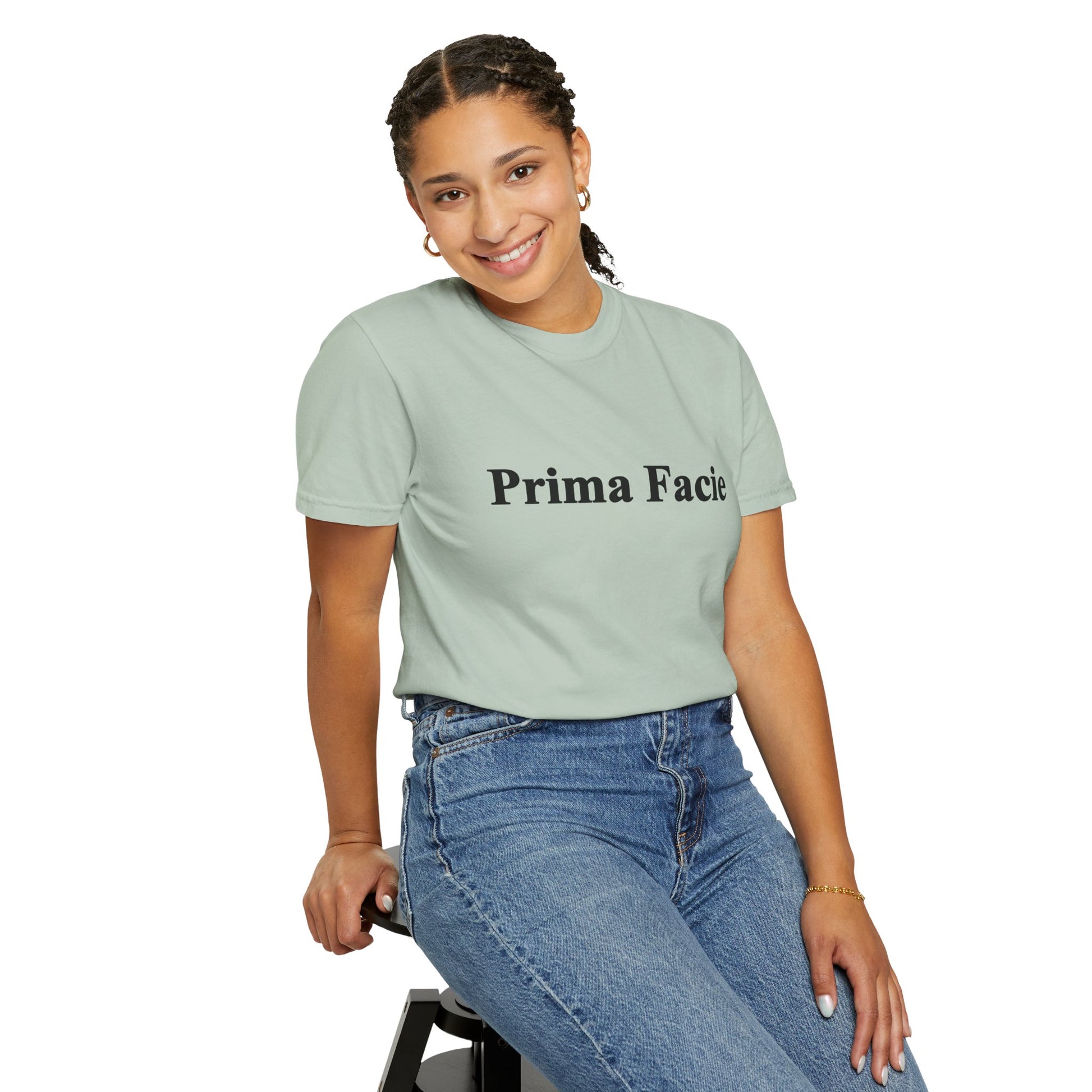 Young female model seated casually while wearing a mint Prima Facie unisex graphic T-shirt in a relaxed studio portrait.
