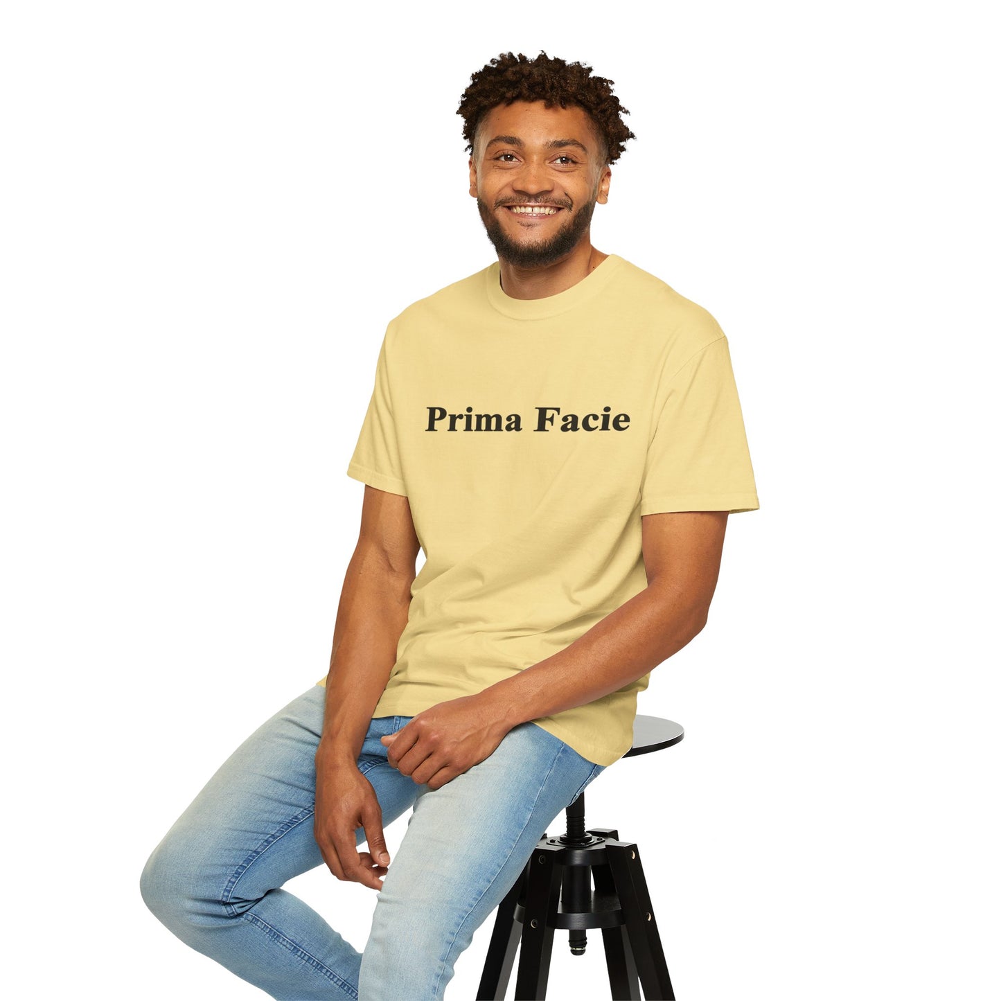 Young male model seated on a stool wearing a pale yellow Prima Facie unisex graphic T-shirt in a casual studio portrait.