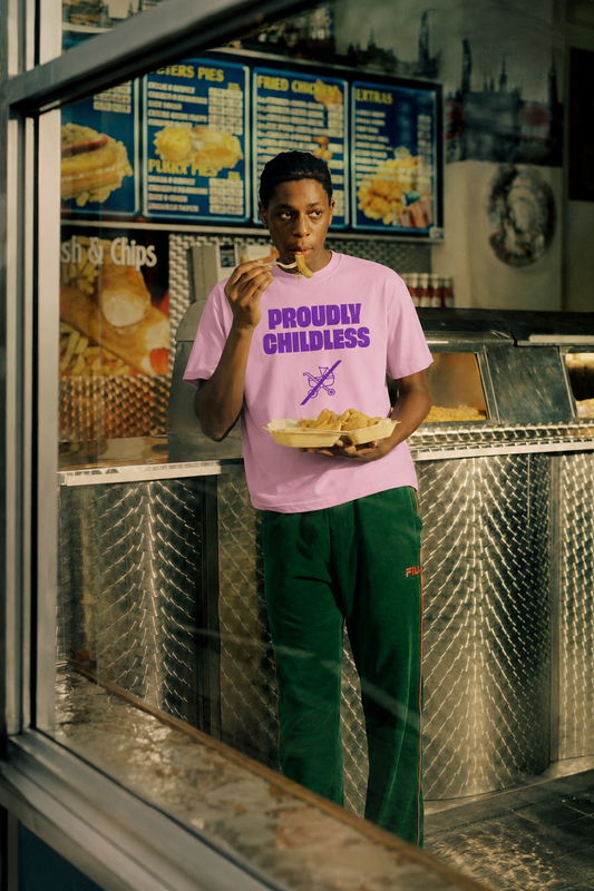 Male model wearing a pink Proudly Childless graphic T-shirt inside a retro fish-and-chips shop