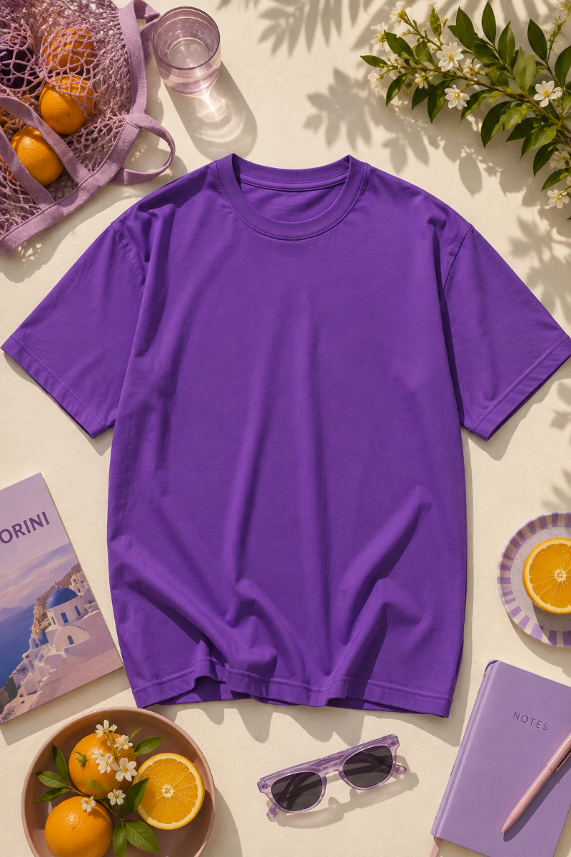 Purple short-sleeve T-shirt flat lay on a light beige surface with oranges, sunglasses, flowers, notebook and summer lifestyle accessories.