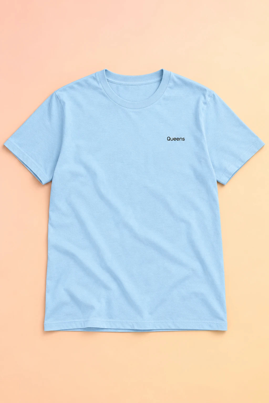 Light blue Queens T-shirt laid flat on a soft peach gradient background, showing small black chest lettering.