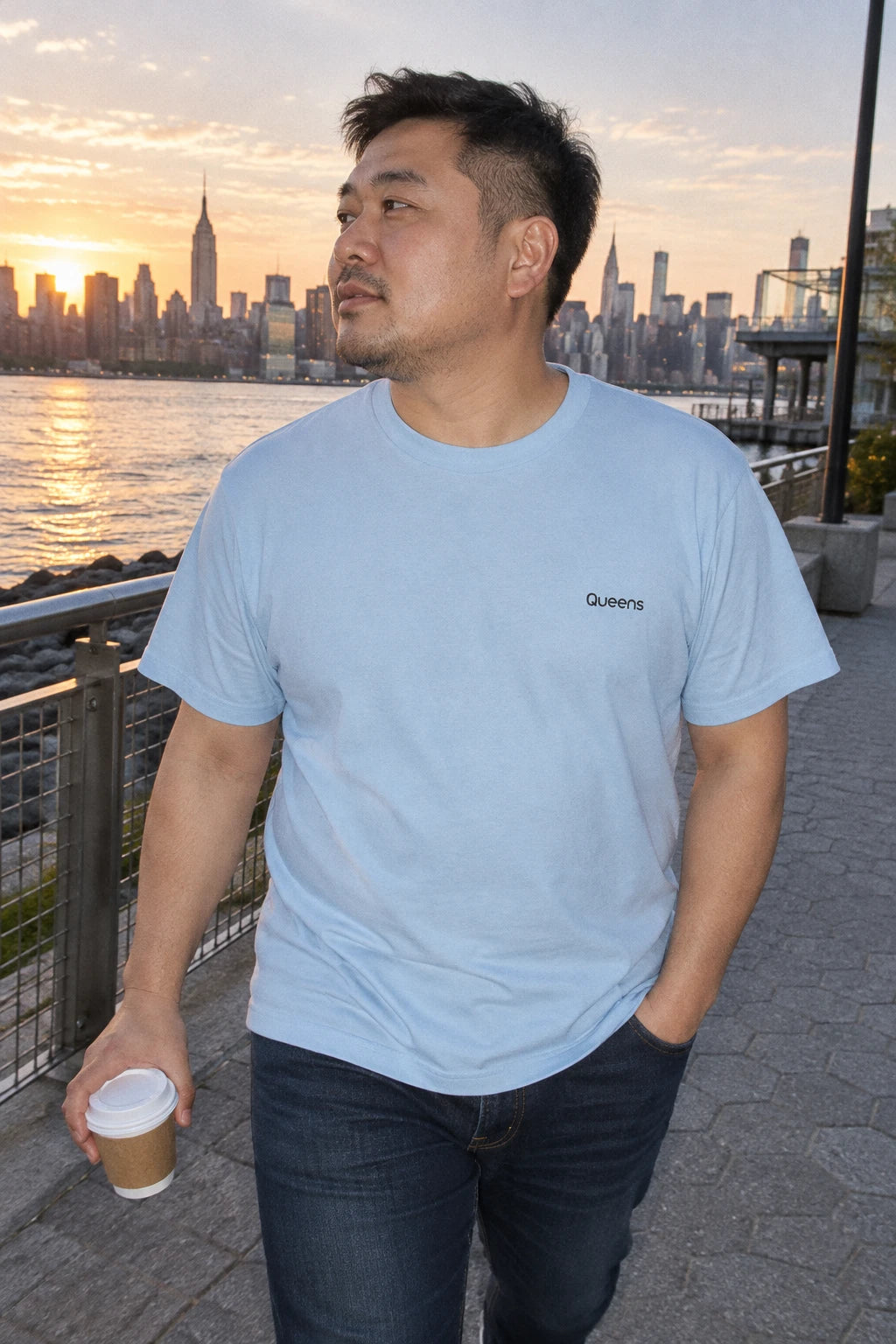 Man wearing a light blue Queens T-shirt while holding coffee beside the waterfront with the Manhattan skyline at sunset.