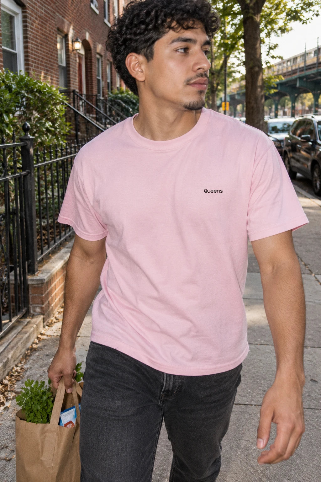 Man wearing a pink Queens T-shirt while carrying groceries along a tree-lined neighborhood sidewalk.