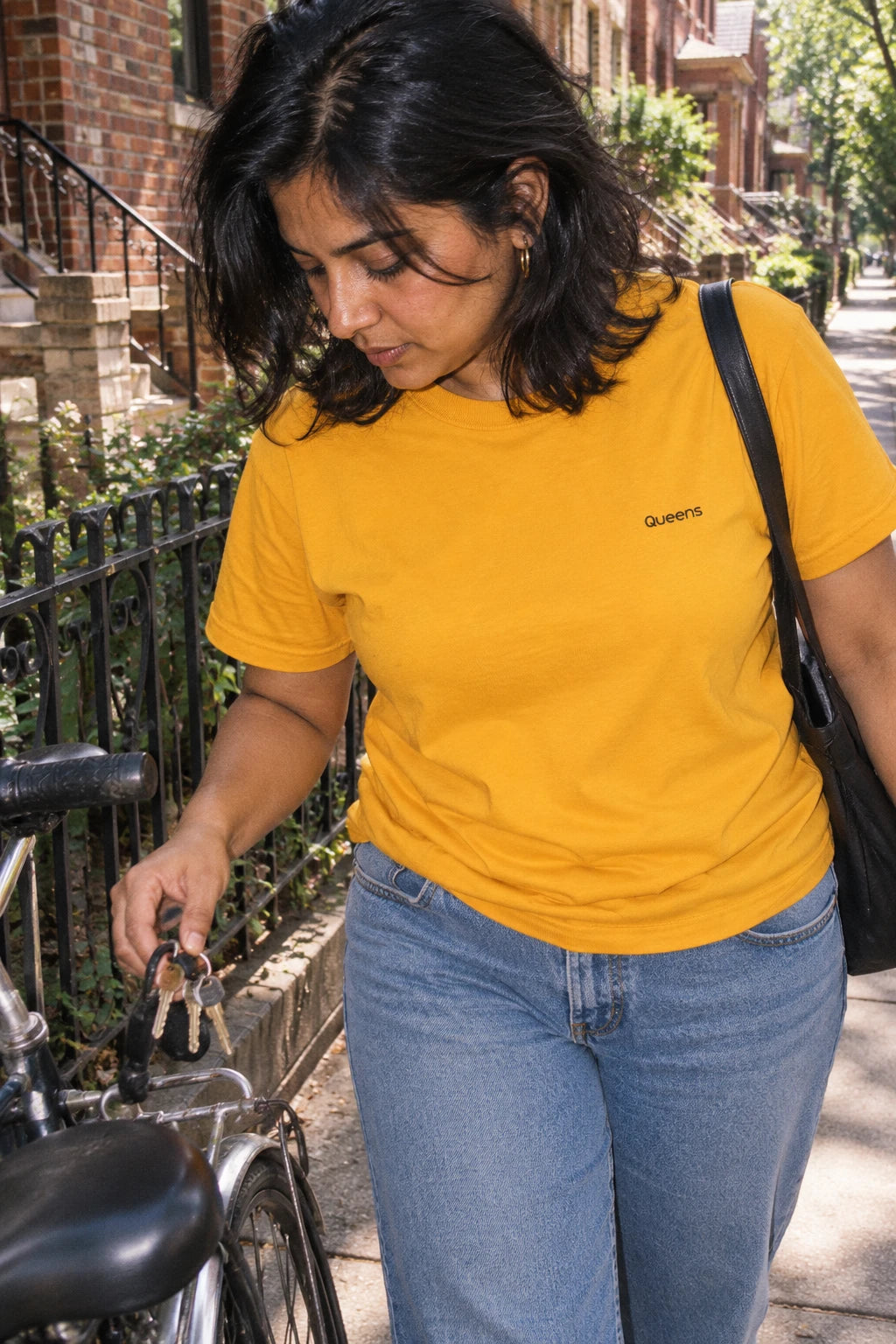 Woman wearing a yellow Queens T-shirt while holding keys beside a bicycle on a leafy neighborhood sidewalk.