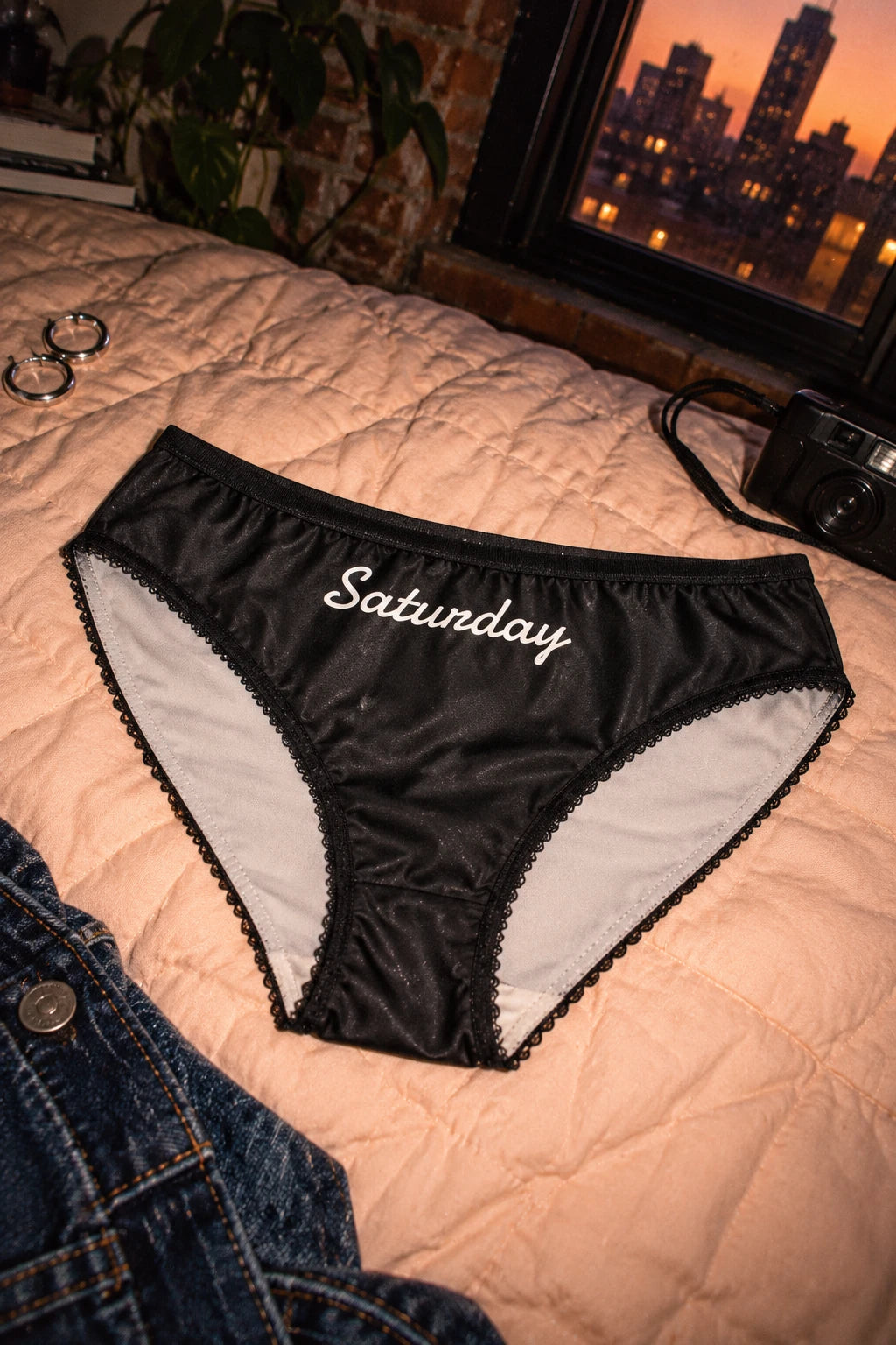 Tantrum Media Saturday Panty in black with white script, resting on peach bedding beside a camera and city apartment window at sunset.
