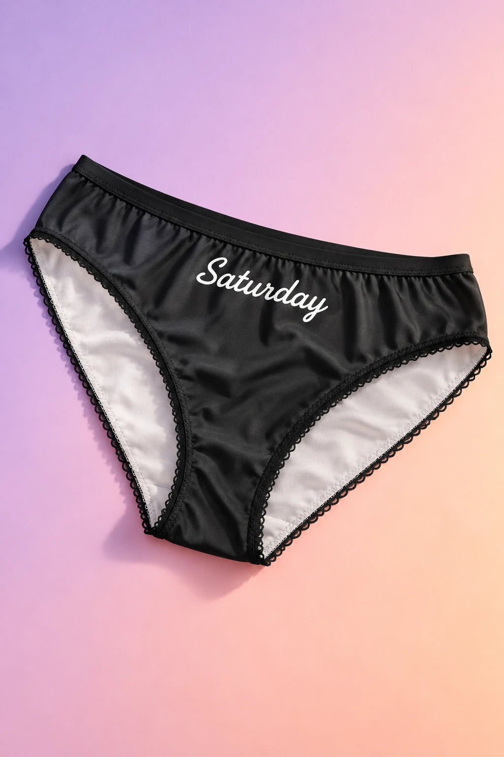 Tantrum Media Saturday Panty in black with white Saturday script and scalloped trim, laid flat on a lavender and peach gradient background.