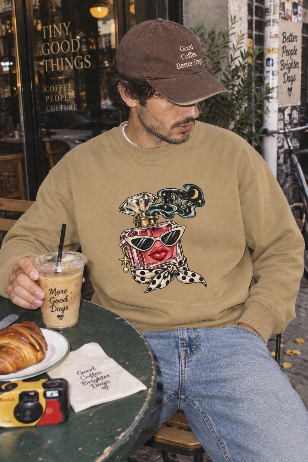 Man wearing the Scent of Chaos tan heavyweight graphic sweatshirt at an outdoor urban café, featuring original surreal perfume bottle artwork.