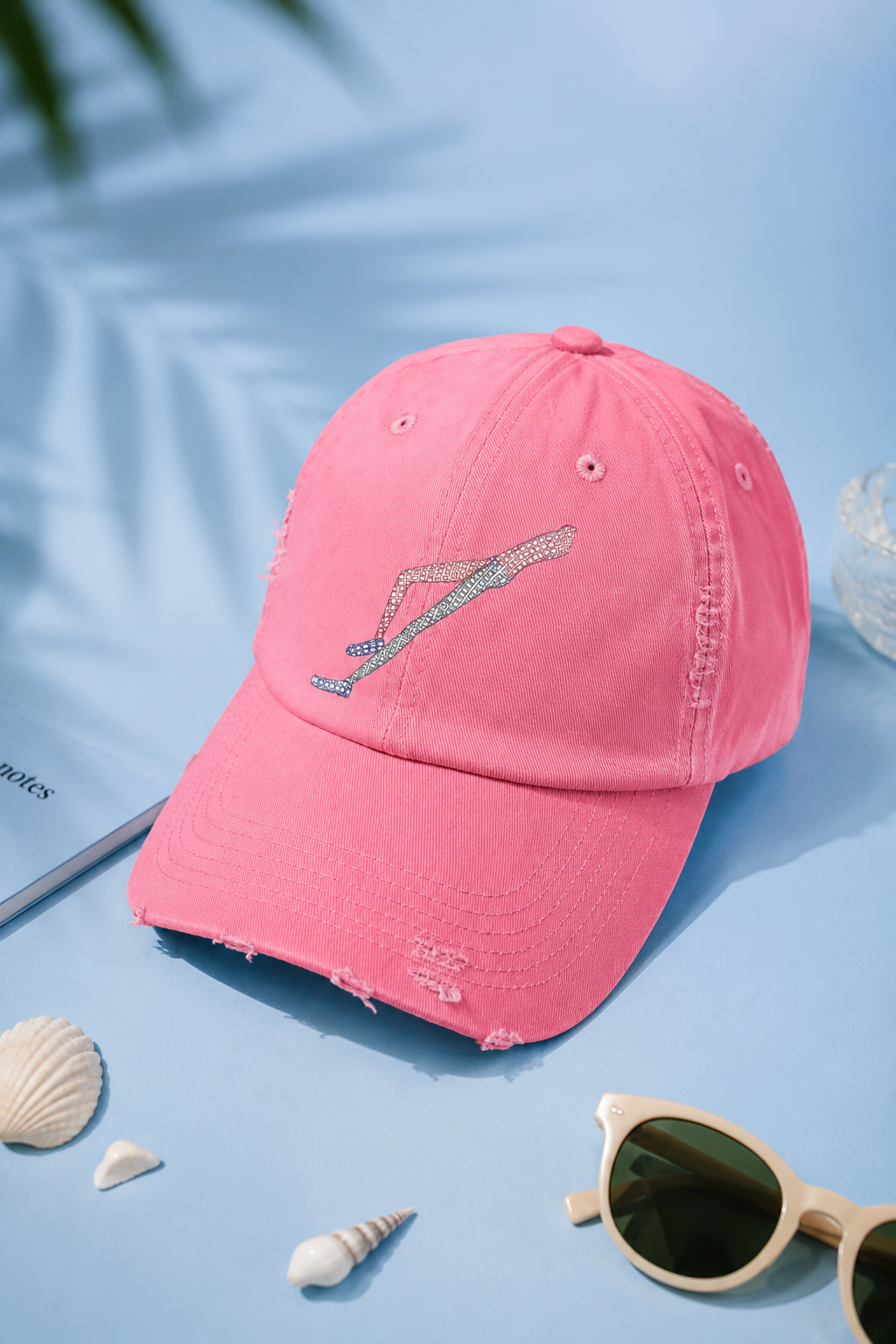 Shapes in Shapes pink distressed baseball cap flat lay on a soft pastel blue background with summer accessories.