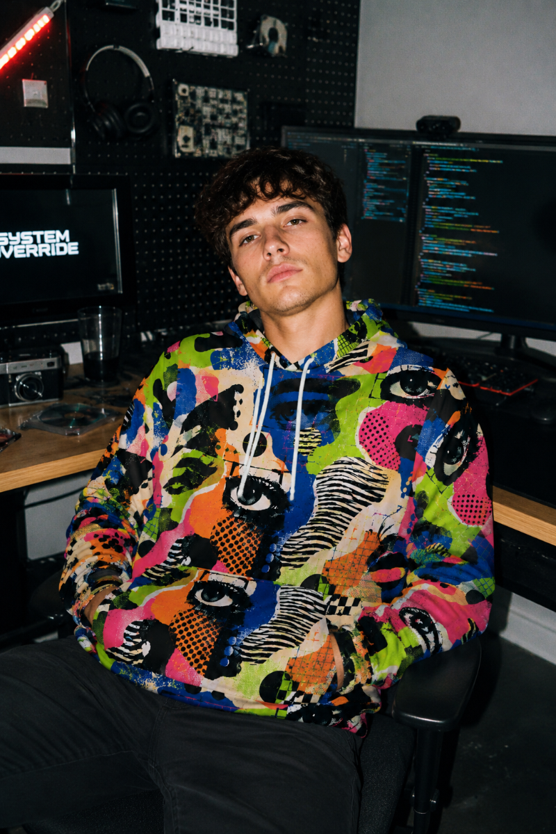 Gen Z man wearing the Signal Jammer adversarial fashion hoodie in a tech workspace with disposable camera aesthetic
