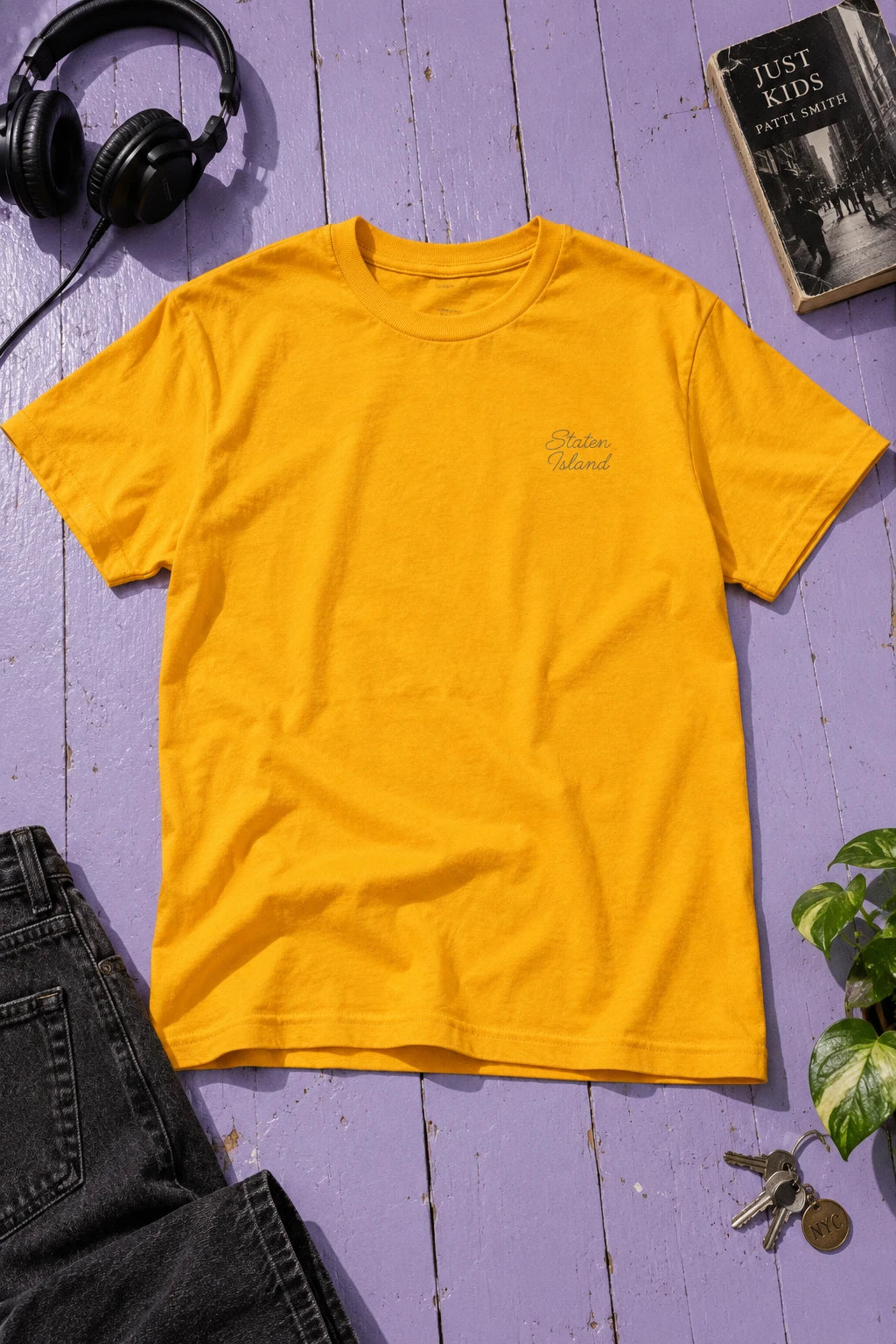 Golden yellow Staten Island T-shirt laid flat on lavender floorboards beside headphones, a book and dark jeans.