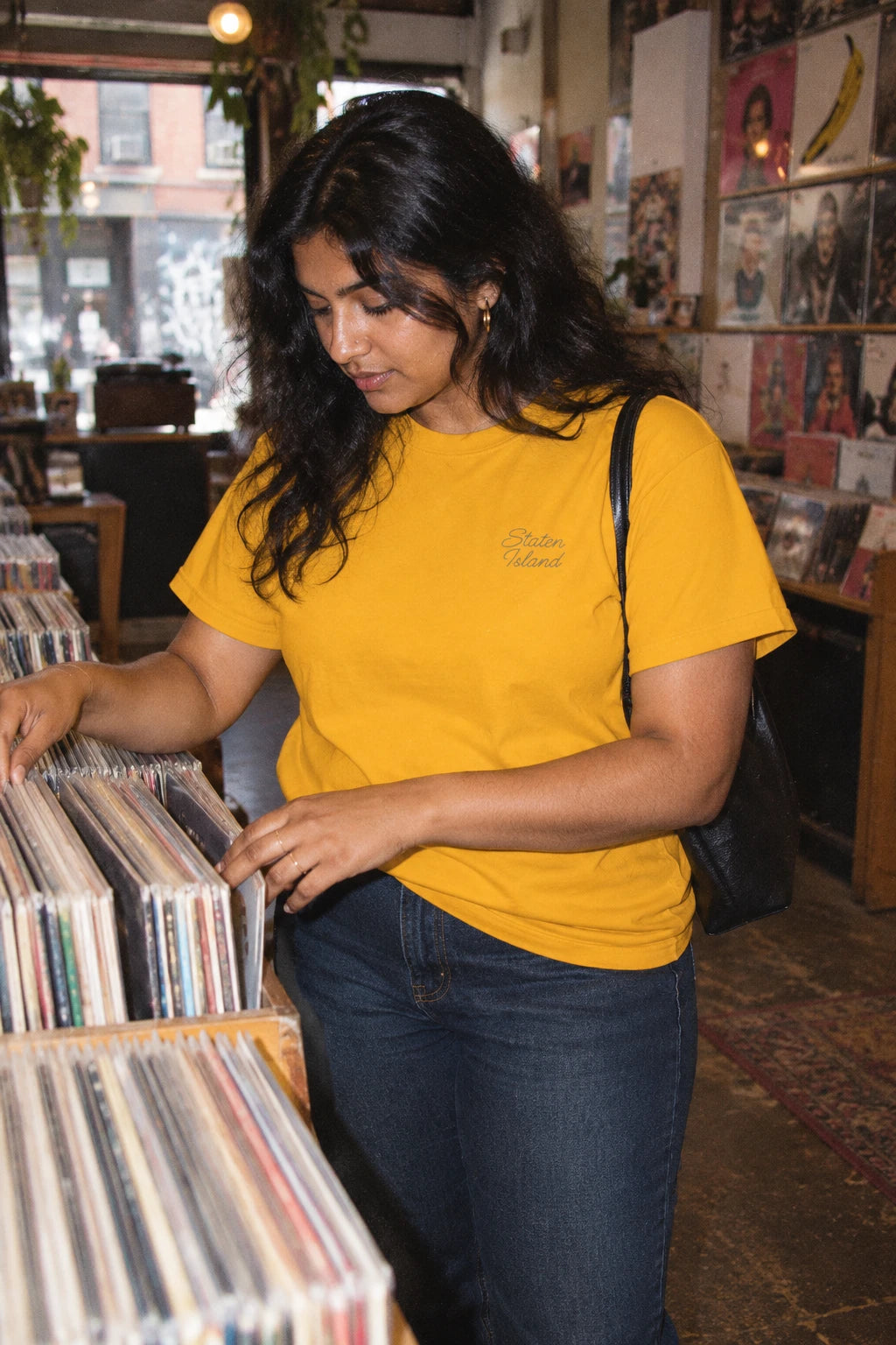 Woman browsing vinyl records while wearing a golden yellow Staten Island script T-shirt.