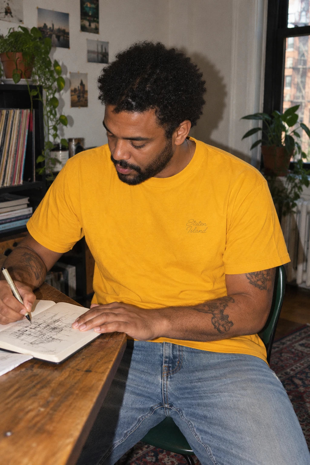 Man wearing a golden yellow Staten Island T-shirt while drawing in a sketchbook at a wooden desk.