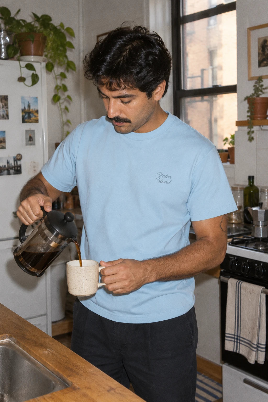 Man wearing a light blue Staten Island T-shirt while pouring coffee in an apartment kitchen.