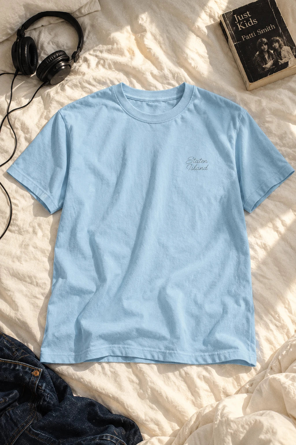 Light blue Staten Island script T-shirt laid flat on ivory bedding beside headphones, a book and dark jeans.