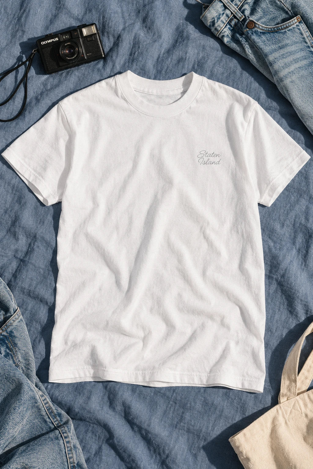 White Staten Island script T-shirt laid flat on blue bedding beside a compact camera and faded jeans.