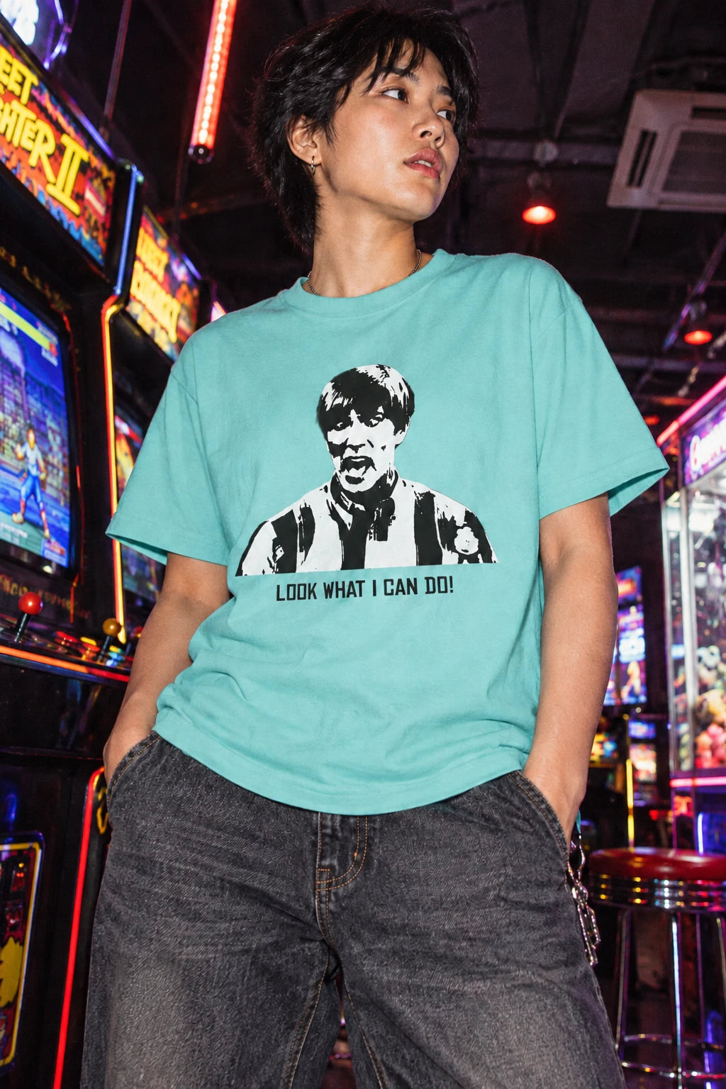 Person wearing the aqua Tantrum Media Stuart MadTV Unisex T-Shirt with dark jeans among illuminated arcade machines.