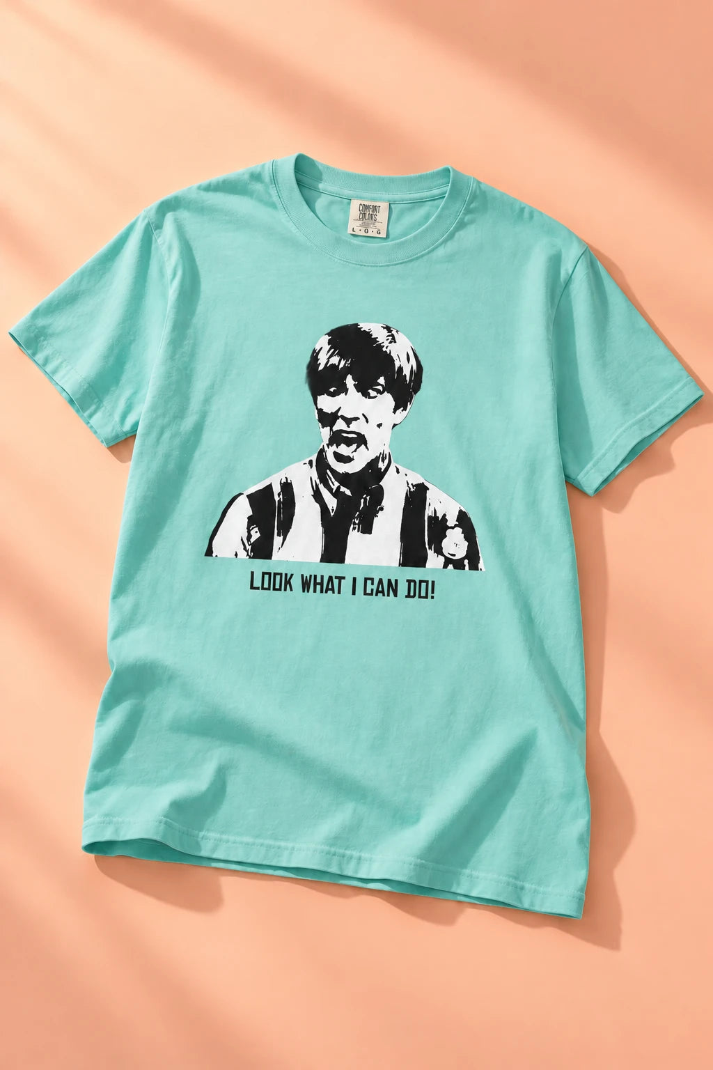 Aqua Tantrum Media Stuart MadTV Unisex T-Shirt laid flat on peach with directional light and soft fabric folds.
