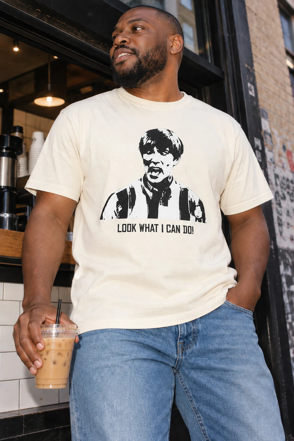 Man wearing the natural white Tantrum Media Stuart MadTV Unisex T-Shirt and jeans, holding iced coffee outside a coffee shop.