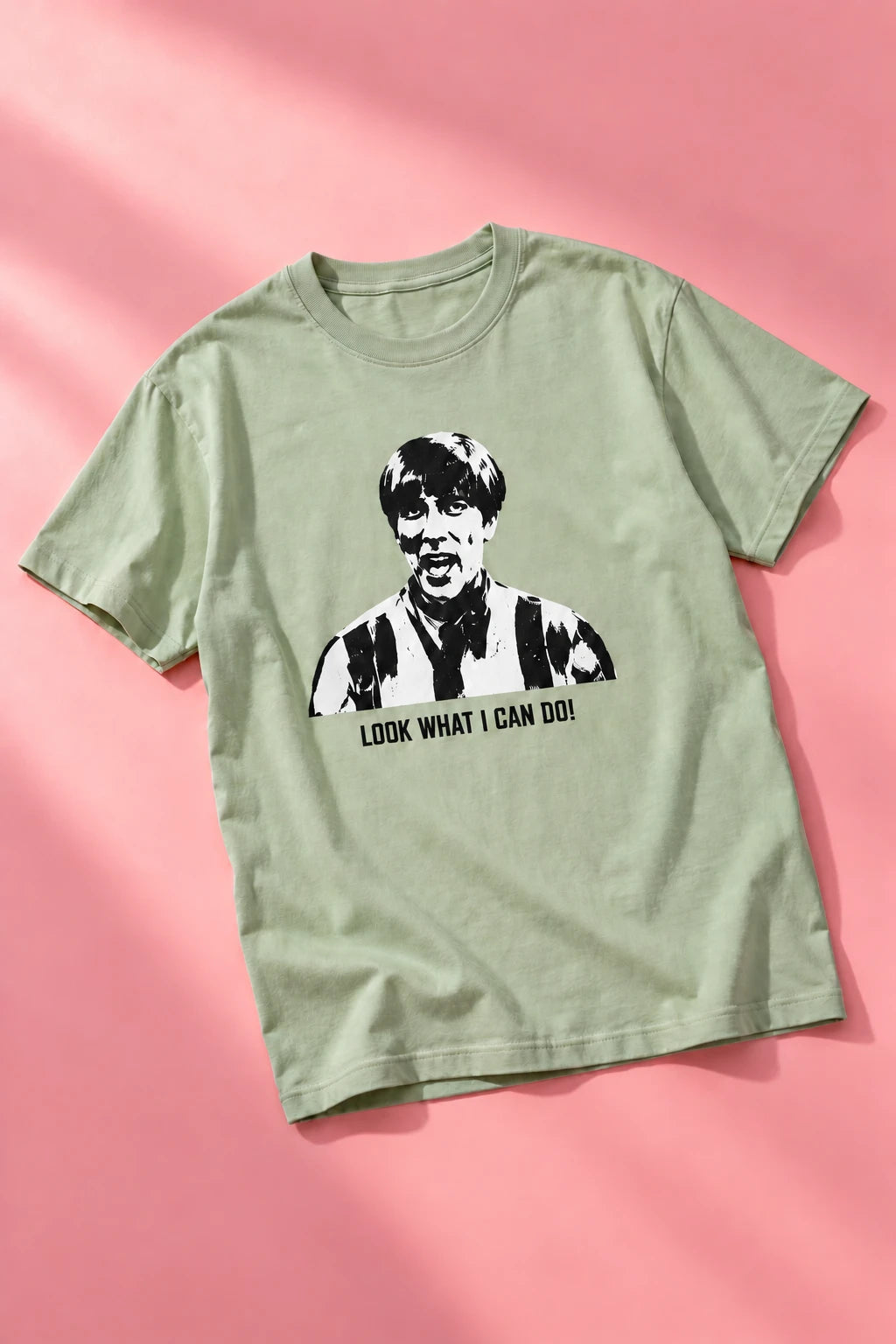 Sage green Tantrum Media Stuart MadTV Unisex T-Shirt laid diagonally on a pink background with soft shadows.