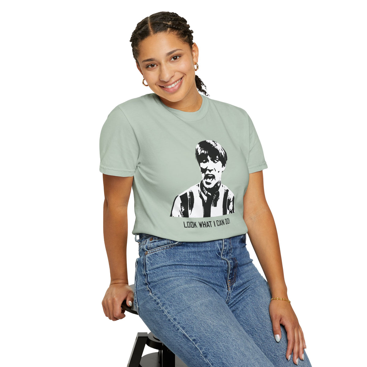 Seated model wearing the sage green Tantrum Media Stuart MadTV Unisex T-Shirt with blue jeans against a white background.