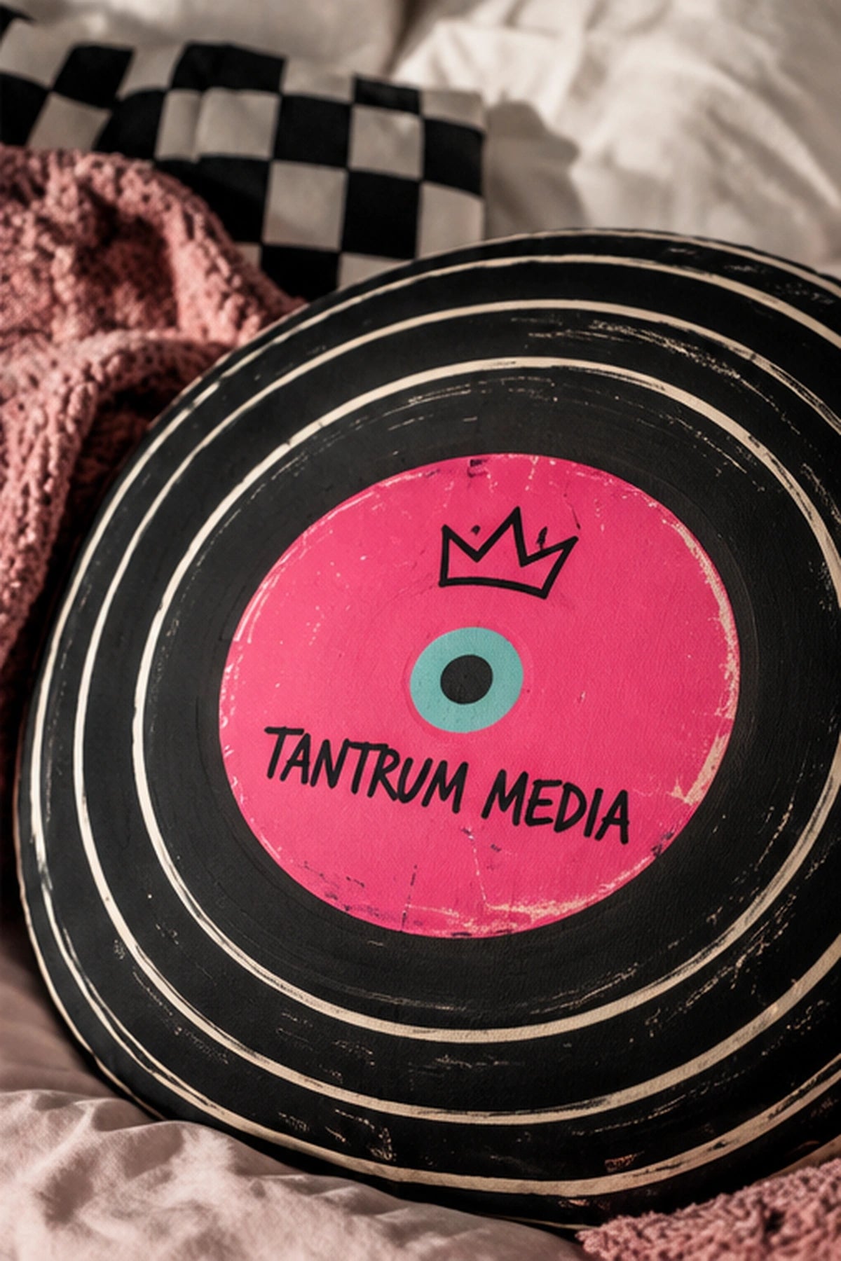 Tantrum Media LP Record Pillow displayed on soft bedding with a checkered cushion and pink blanket, featuring black vinyl grooves and a pink record label.