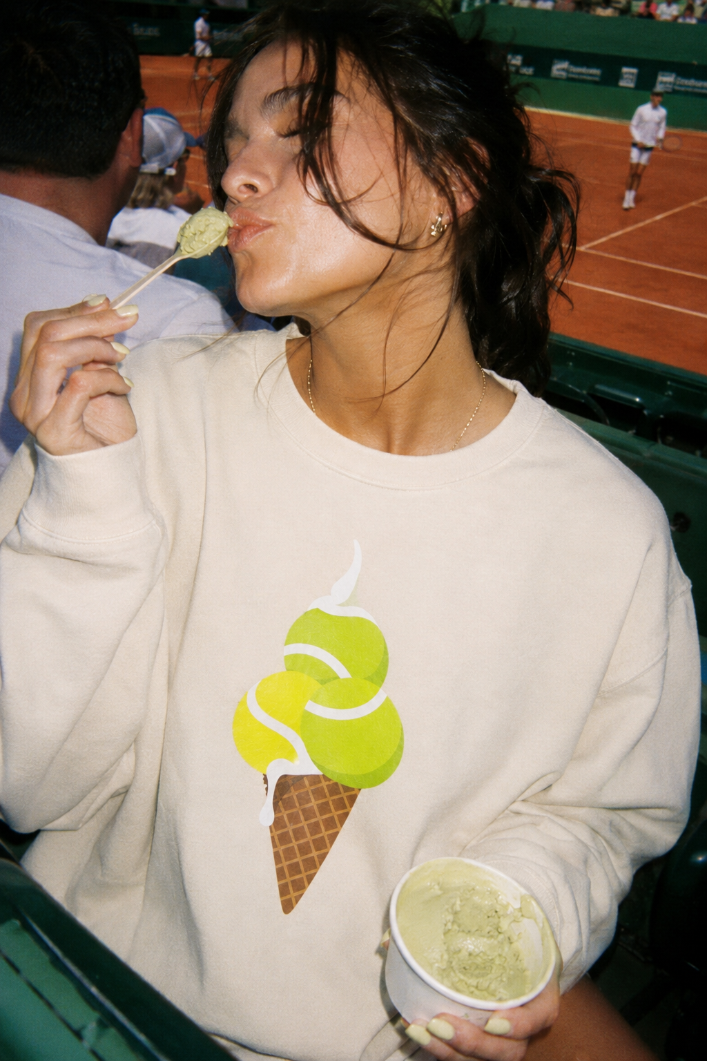 Woman in cream tennis sweatshirt eating pistachio ice cream courtside at a tennis match