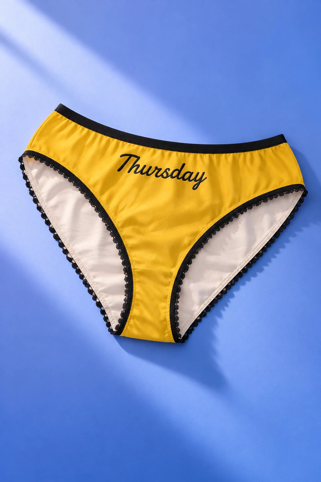 Tantrum Media Thursday Panty in yellow with black Thursday script and scalloped trim, laid flat on a blue gradient background.