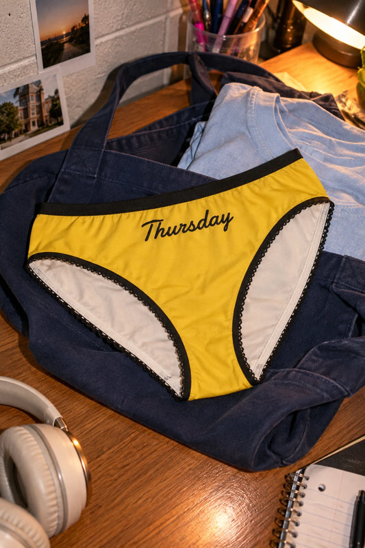 Tantrum Media Thursday Panty in yellow with black script and trim, resting on a navy tote beside headphones on a dorm desk.