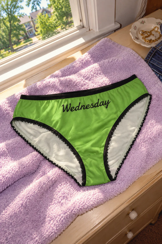 Tantrum Media Wednesday Panty in green with black script and trim, resting on a lavender towel beside a sunlit bedroom window.