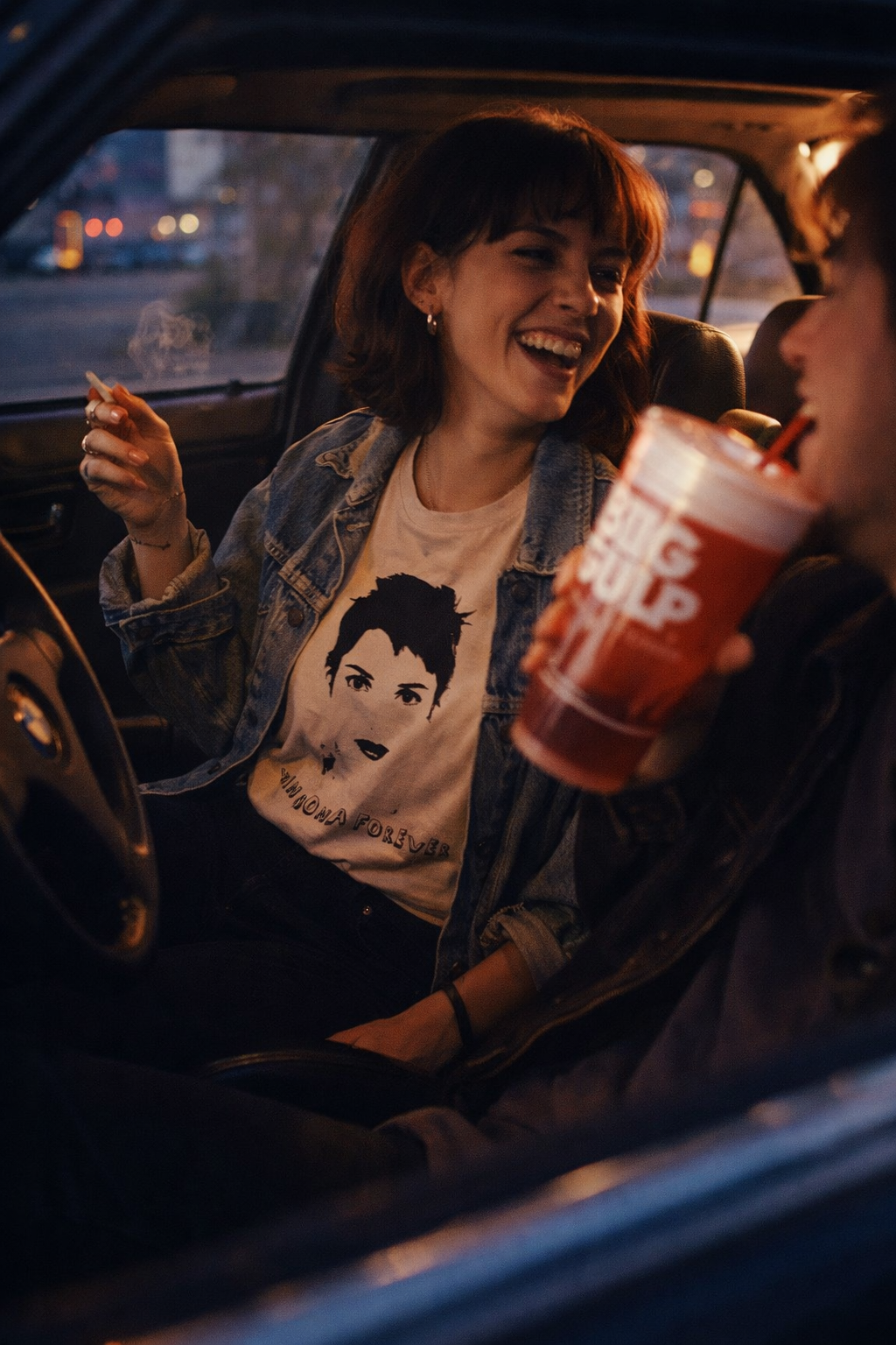 Winona Forever T-Shirt cream graphic tee worn by a woman enjoying a drink inside a car at night
