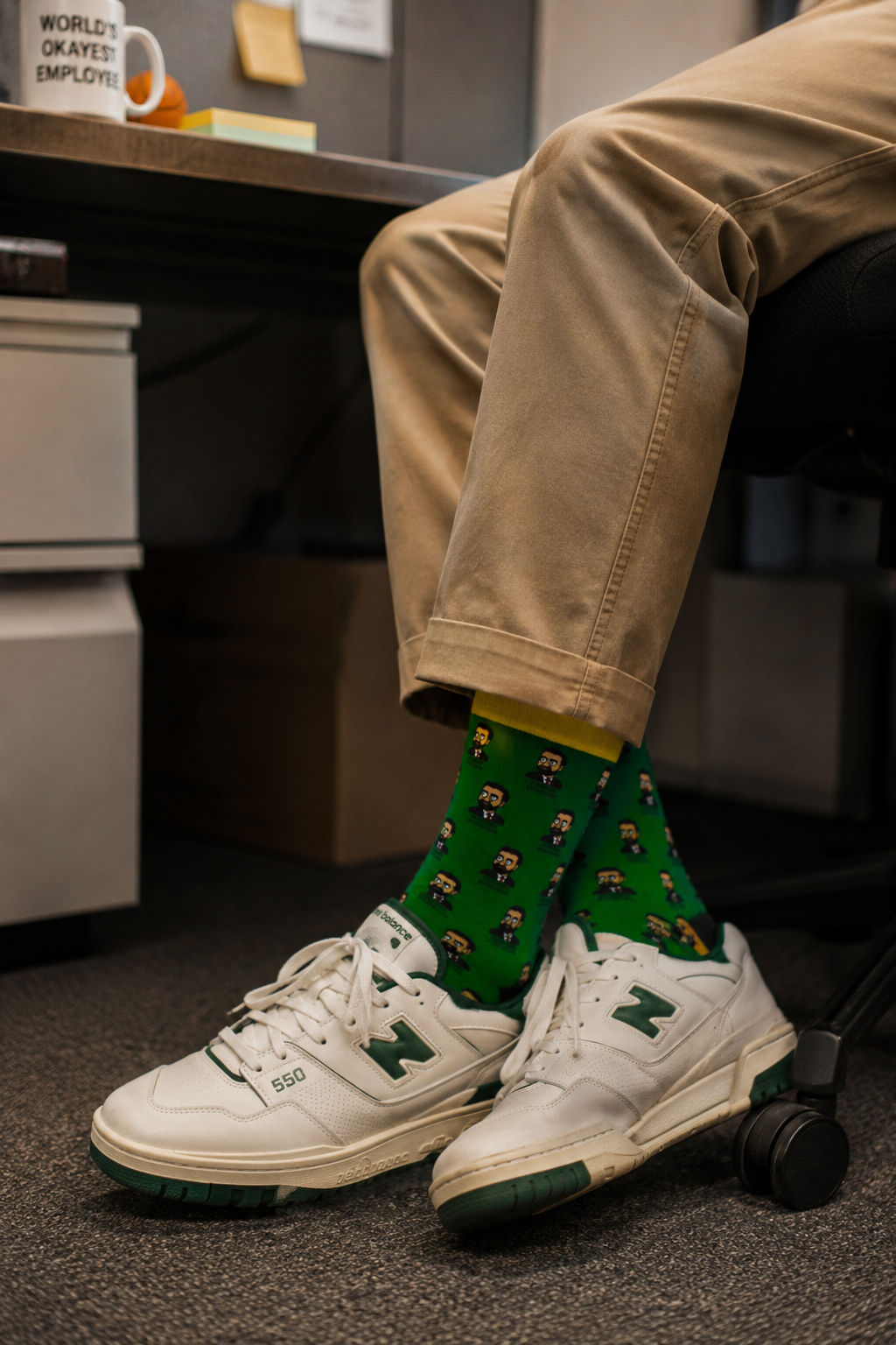 Male Gen Z professional wearing Zohran Mamdani unisex padded crew socks with white sneakers while sitting in a modern office cubicle, showcasing green novelty socks with repeating Zohran Mamdani artwork.